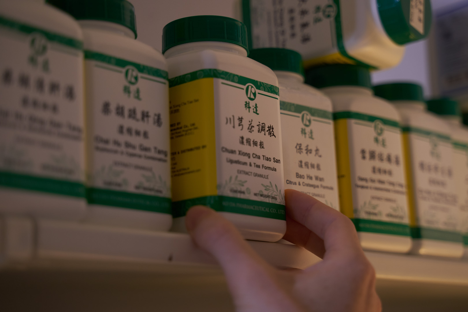 A hand selecting a bottle of Chinese herbal medicine from a shelf with similar bottles.