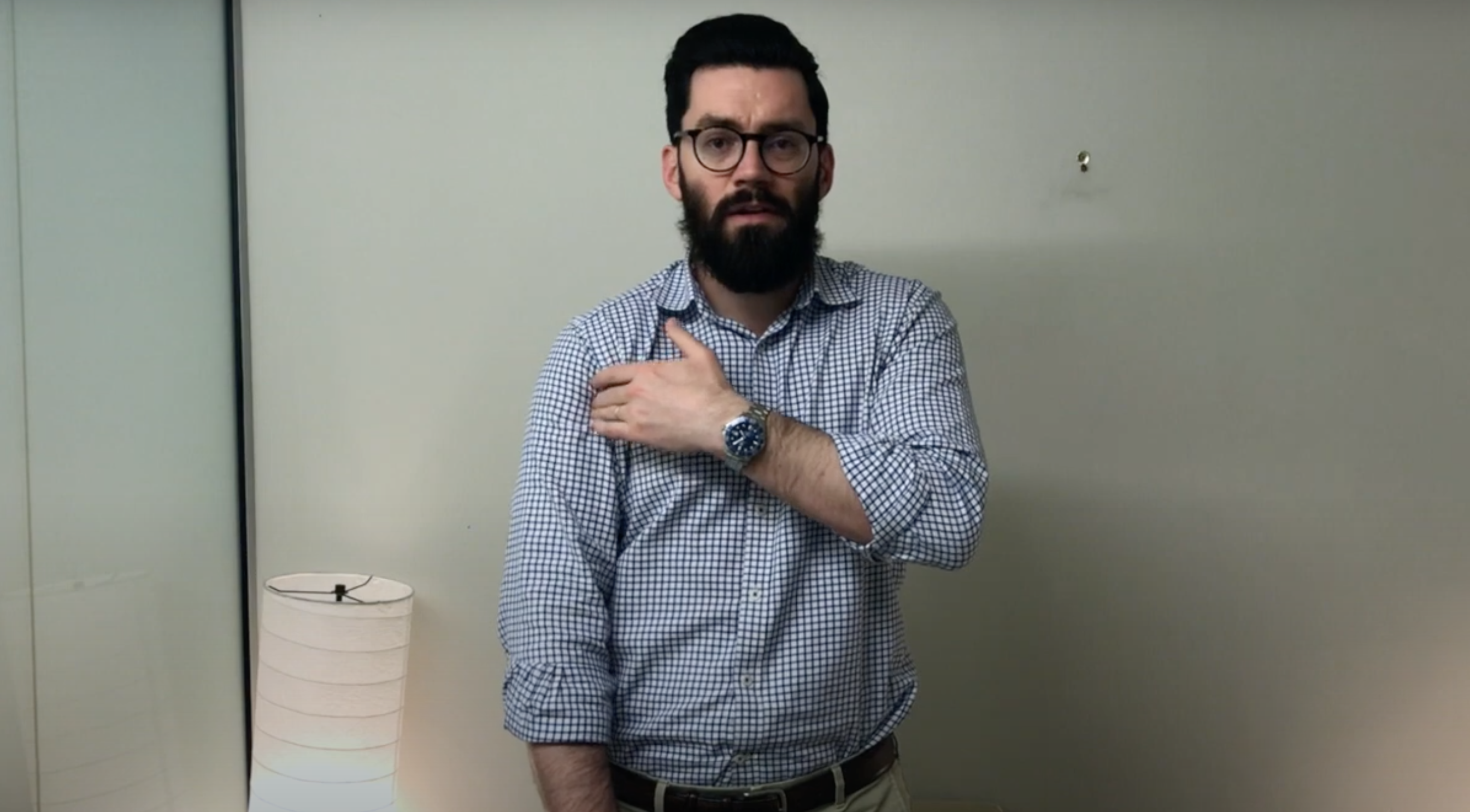Man in glasses touches his left shoulder, suggesting TCM or acupuncture near a blank wall indoors.